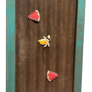 May include: A rustic, turquoise-framed board with a corrugated metal backing. Three colorful magnets are attached: two watermelon slices and a banana wearing sunglasses. The board is designed for displaying notes or photos.