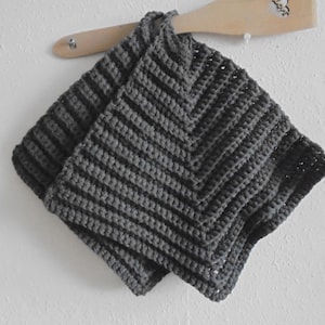 May include: Two dark grey, handmade crochet potholders with a ribbed chevron pattern are hanging on a wooden spatula. The potholders are square and folded, showcasing the textured stitch work. The background is a plain white wall.