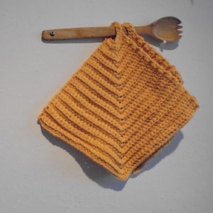 May include: An orange crocheted potholder with a chevron pattern hangs from a wooden spoon on a light grey wall. The potholder is square and has a textured appearance, with visible stitch lines creating the design.