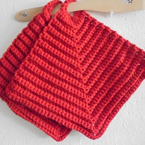 Könnte beinhalten: Zwei rote gehäkelte Topflappen in quadratischer Form mit einem strukturierten Muster. Die Topflappen haben oben eine Schlaufe zum Aufhängen.