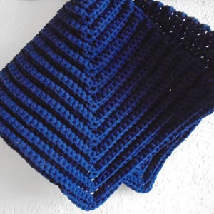 Op de afbeelding: Een gevouwen, handgemaakte gehaakte deken in blauwtinten. De deken heeft een geribbeld patroon met een geschulpte rand. Het garen lijkt zacht en de deken is netjes opgevouwen, wat de textuur en het ontwerp laat zien.