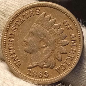 Puede incluir: Una moneda de cobre de 1863, Indian Head. La moneda muestra el perfil de un nativo americano con un tocado de plumas, con las palabras "UNITED STATES OF AMERICA". El año "1863" es visible en la parte inferior.