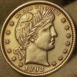 May include: A gold-colored 1892 half dollar coin with the words "In God We Trust" and "Liberty" inscribed on it. The coin features a profile of a woman with a wreath on her head.