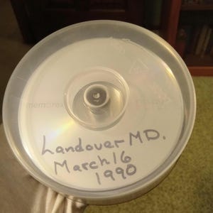 May include: A stack of blank CDs in a clear plastic case. The CDs are white with a silver reflective surface. The case has the text "Landover MD. March 16 1990" written on it.
