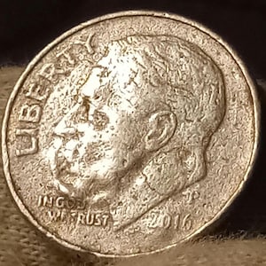 May include: A close-up of a silver United States dime coin. The coin features a profile of a man's head, the word "LIBERTY" at the top, and the words "IN GOD WE TRUST" and the date "2016" at the bottom.