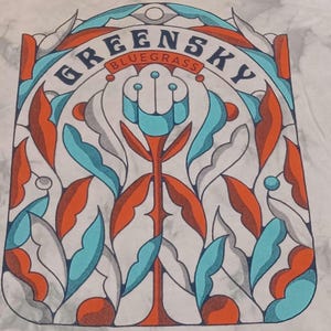 May include: White t-shirt featuring a colorful stained-glass style design. The design includes the words "Greensky Bluegrass" in an arch, with orange, blue, and gray leaf-like shapes. The overall aesthetic is reminiscent of a vintage poster.