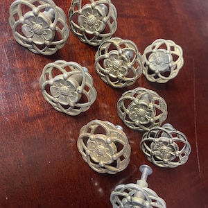 May include: A selection of antique cabinet knobs. Each knob has a floral design at the centre, enclosed by an openwork, cage-like structure. The knobs are made of a silver-toned metal and are arranged on a dark wood surface.