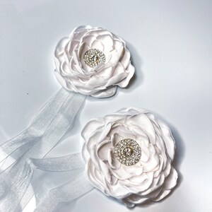 以下が含まれることがあります： ラインストーンのセンターが付いた白いサテン生地の花が2つ。それぞれの花は白いリボンに付けられています。
