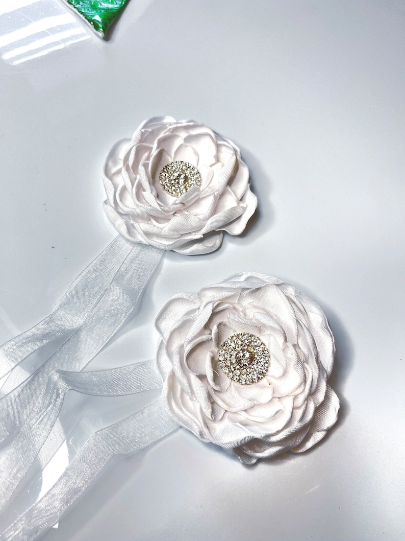 以下が含まれることがあります： ラインストーンのセンターが付いた白いサテン生地の花が2つ。それぞれの花は白いリボンに付けられています。