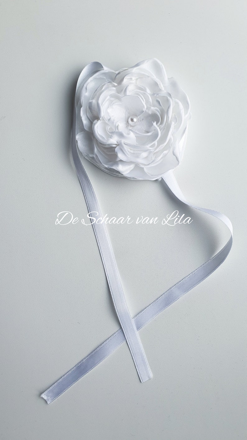 以下が含まれることがあります： 白いサテンのリボンにパールの中心を持つ白いサテンの花が取り付けられています。リボンには「De Schaar van Lila」と印刷されています。