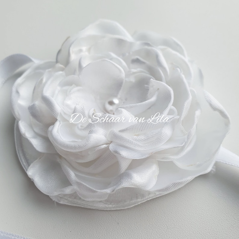 以下が含まれることがあります： 真珠の中心を持つ白いサテンの花。花は白いリボンに付けられています。花には「De Schaar van Lila」と書かれています。