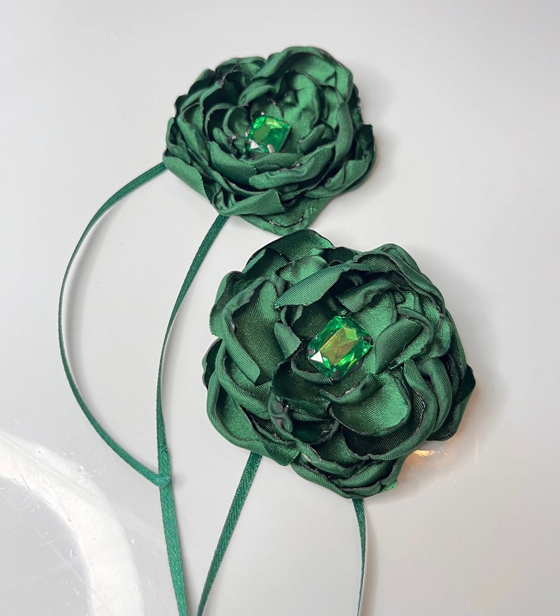 以下が含まれることがあります： 緑色の宝石が中央に付いた、濃い緑色の布製のフラワーブローチが2つ