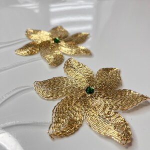 以下が含まれることがあります： 緑色の宝石の中心を持つ、金色のビーズでできた花の形をした装飾品が2つ。花は白い表面の上に置かれています。