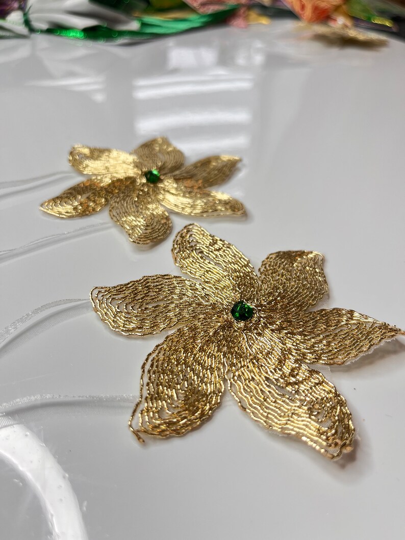 以下が含まれることがあります： 緑色の宝石の中心を持つ、金色のビーズでできた花の形をした装飾品が2つ。花は白い表面の上に置かれています。