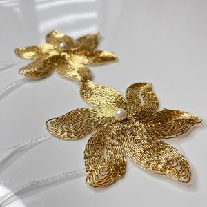 以下が含まれることがあります： 白い真珠のアクセントが付いた、金色のビーズでできた花の形が2つ。花は白いリボンで繋がっています。