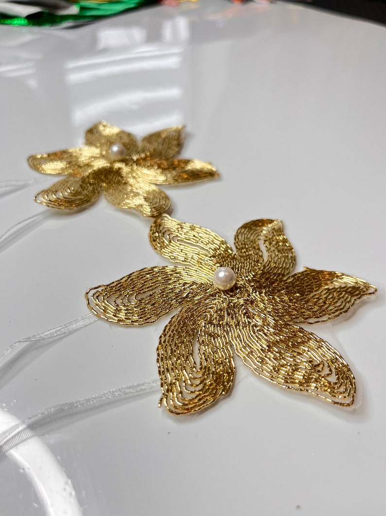 以下が含まれることがあります： 白い真珠のアクセントが付いた、金色のビーズでできた花の形が2つ。花は白いリボンで繋がっています。