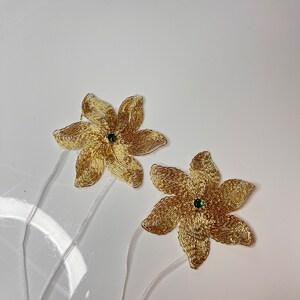 以下が含まれることがあります： 緑色の宝石の中心を持つ、金色のビーズでできた花の装飾が2つ。花は白いリボンで茎につけられています。