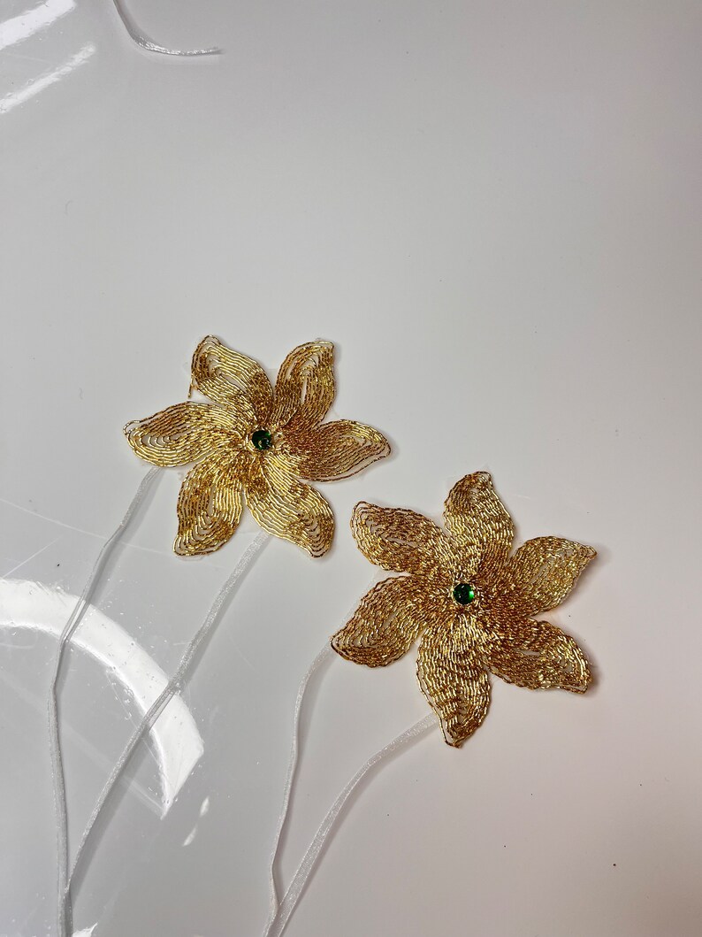 以下が含まれることがあります： 緑色の宝石の中心を持つ、金色のビーズでできた花の装飾が2つ。花は白いリボンで茎につけられています。