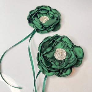 以下が含まれることがあります： ラインストーンのセンターが付いた、濃い緑色の布製の2つの花。花は緑のリボンに付けられています。