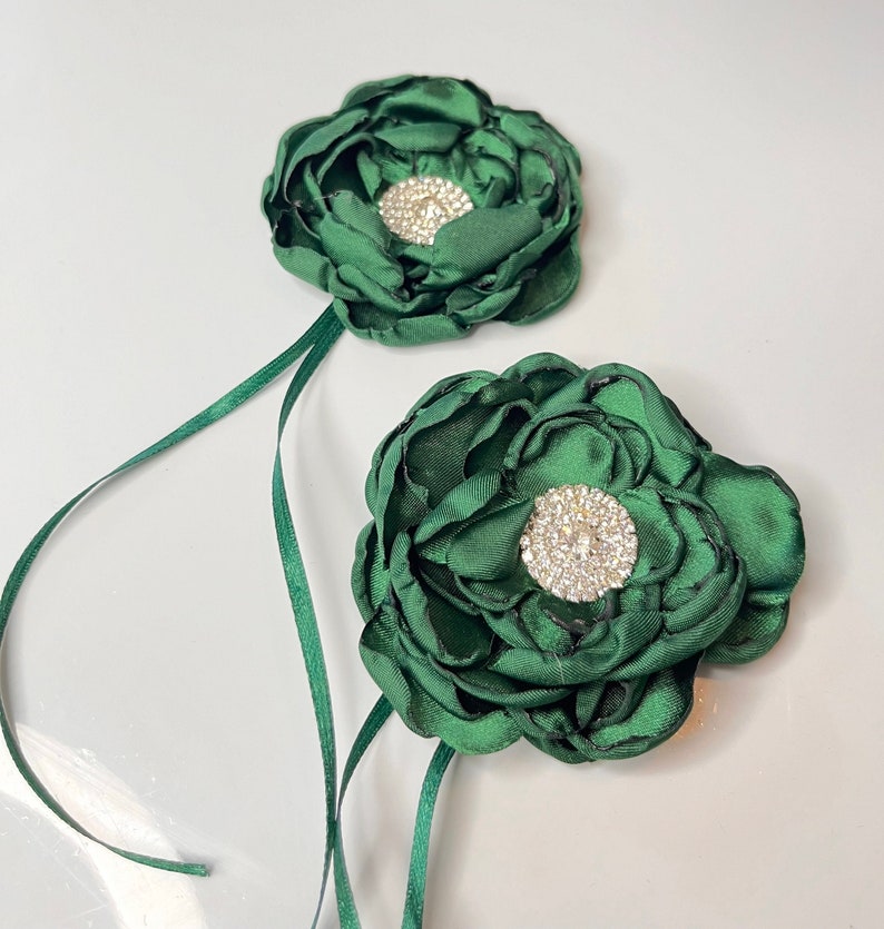 以下が含まれることがあります： ラインストーンのセンターが付いた、濃い緑色の布製の2つの花。花は緑のリボンに付けられています。