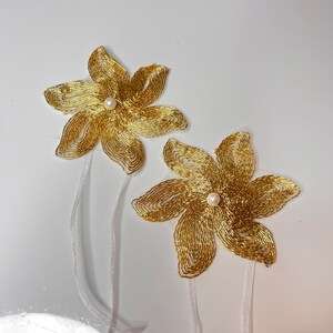 以下が含まれることがあります： 白い真珠の中心と白いサテンリボンタイが付いた、金色のビーズでできた花の装飾が2つです。