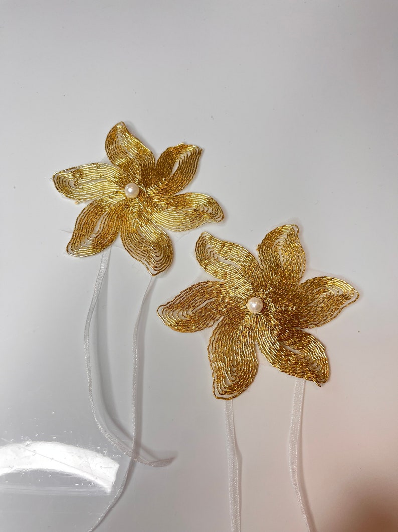 以下が含まれることがあります： 白い真珠の中心と白いサテンリボンタイが付いた、金色のビーズでできた花の装飾が2つです。