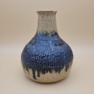 May include: A ceramic vase with a rounded body and a narrow neck. The vase is glazed in a light beige color on the top half and a dark blue color on the bottom half. The blue glaze has a textured, drippy effect.