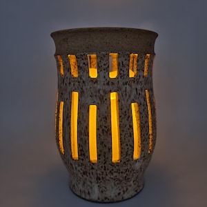 May include: A ceramic lantern with vertical rectangular cutouts. The lantern has a speckled brown and cream exterior and a tan rim. The interior glows with a warm yellow light, creating a cozy ambiance. This decorative piece is ideal for home decor.