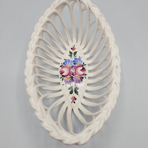 May include: White ceramic oval basket with an openwork design. The basket features a braided edge and a painted floral motif in the center with pink, purple, and blue flowers. A decorative piece for the home.