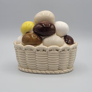May include: A ceramic basket filled with a variety of colorful eggs. The basket is white with a woven pattern and has a scalloped edge. The eggs are white, brown, and yellow.