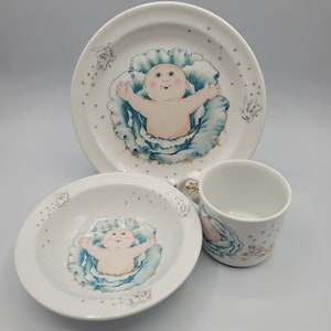 May include: A white porcelain children's dinnerware set featuring a blue cabbage and a baby design. The set includes a plate, bowl, and cup.
