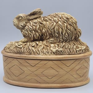 May include: A beige ceramic jewelry box with a detailed rabbit figurine on the lid. The rabbit is lying on a bed of grass with small flowers. The box has a diamond pattern around the sides.