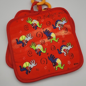 May include: A square red pot holder featuring a repeating pattern of Kokopelli figures in green, blue, and yellow. The word Arizona is printed at the top, and the letters SSI appear in the bottom right corner. An orange plastic ring is attached.