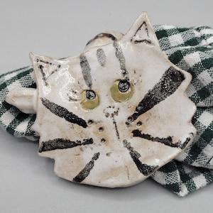 May include: A handmade ceramic cat face, painted white with black stripes and green eyes. The cat-shaped object is resting on a green and white checkered cloth. The cat has a whimsical, handcrafted appearance.