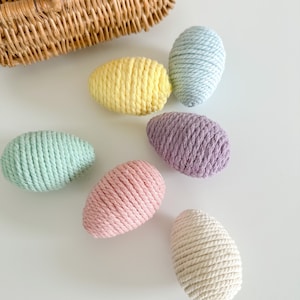 Pode incluir: Seis ovos de Páscoa coloridos envoltos em fio de lã em cores pastel: azul claro, amarelo, verde, rosa, roxo e branco. Os ovos estão sobre uma superfície branca.