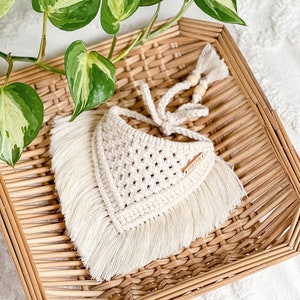 May include: A white crocheted bandana with fringe, tied with a white cord and wooden beads. The bandana has a small brown leather tag with the text "IVORY LANE" on it. The bandana is sitting in a woven wicker basket.