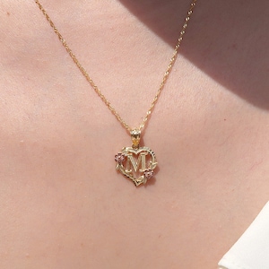 May include: A gold chain necklace with a heart-shaped pendant featuring the letter "M" and two small pink gemstones.