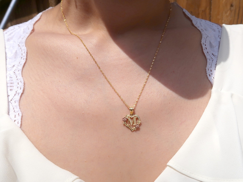 May include: A gold chain necklace with a heart-shaped pendant featuring the letter "M" and two small pink gemstones.