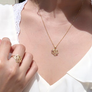 May include: A gold necklace with a heart-shaped pendant featuring the letters "MM" in a decorative font. The pendant has small pink stones around the edges. A matching gold ring with the same design is on the finger of a person's hand.