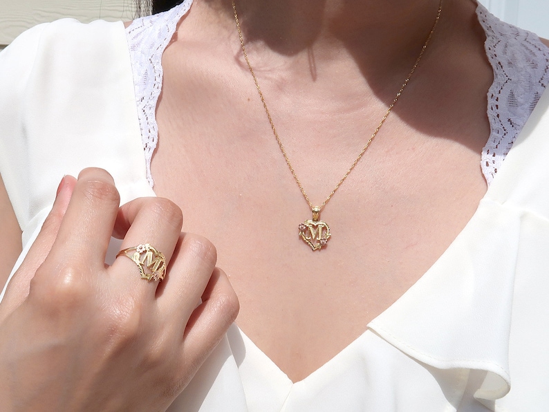 May include: A gold necklace with a heart-shaped pendant featuring the letters "MM" in a decorative font. The pendant has small pink stones around the edges. A matching gold ring with the same design is on the finger of a person's hand.