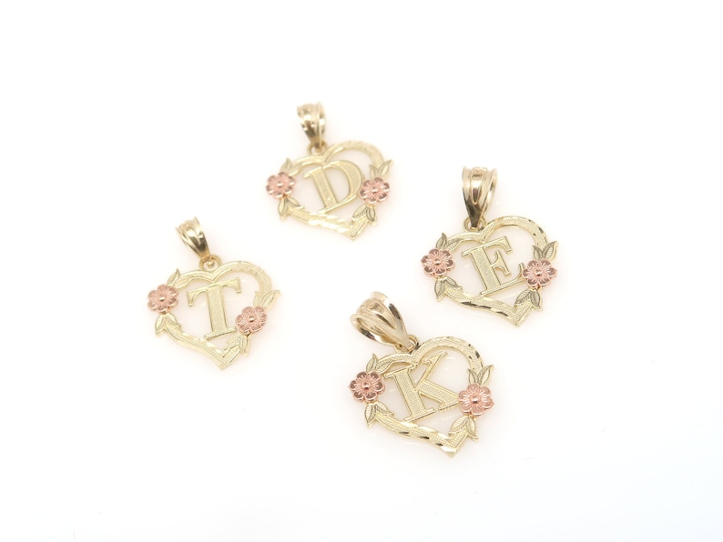 May include: Three gold heart-shaped pendants with different letters, D, E, and K, in the center. Each pendant has a floral design around the letter and pink accents.