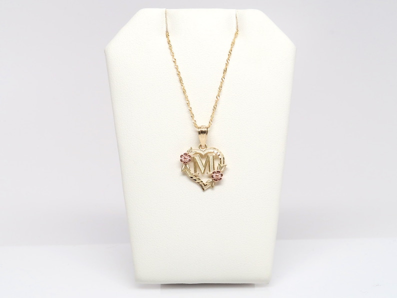 May include: A gold necklace with a heart-shaped pendant featuring the letter "M" surrounded by small pink flowers.