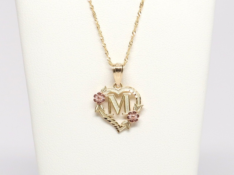 May include: A gold necklace with a heart-shaped pendant featuring the letter "M" surrounded by small pink flowers and leaves.