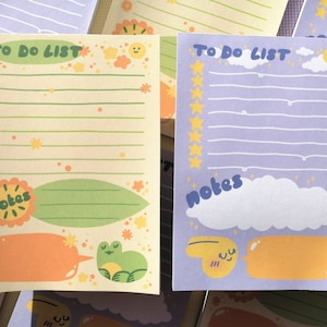 May include: Two rectangular paper to-do list notepads. The left notepad is cream-colored with green lines, floral accents, and a green frog illustration. The right notepad is lavender with blue lines, yellow star bullet points, and a white cloud.
