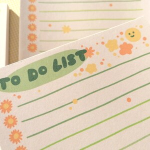 May include: A rectangular notepad with cream-colored paper featuring the text TO DO LIST in dark green letters on a light green background. The page includes horizontal green lines, orange flower icons, and small yellow sun and cloud graphics.