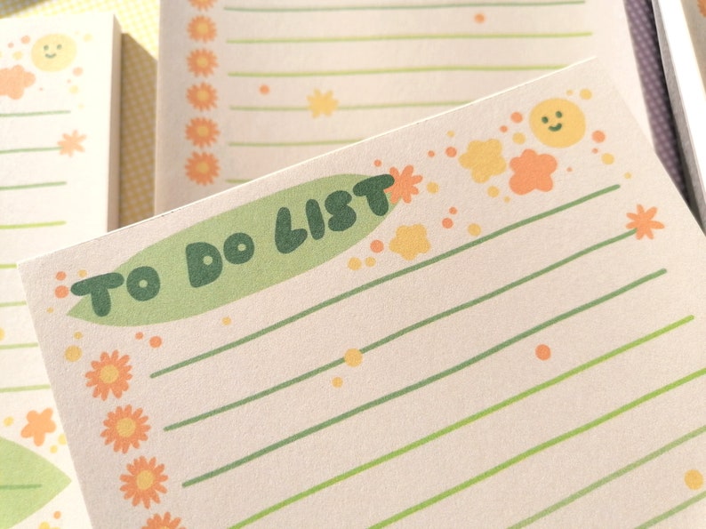 May include: A rectangular notepad with cream-colored paper featuring the text TO DO LIST in dark green letters on a light green background. The page includes horizontal green lines, orange flower icons, and small yellow sun and cloud graphics.