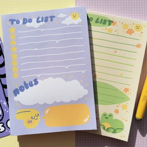 May include: A purple notepad with the text TO DO LIST and NOTES, featuring yellow stars, a sun, clouds, and a smiling yellow character. Beside it is a light green notepad with a frog illustration and a yellow felt-tip pen with Japanese text.