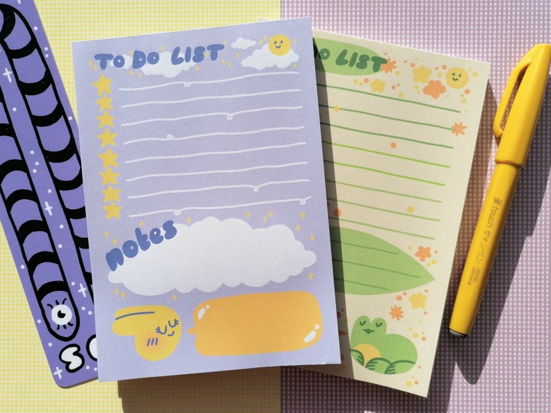 May include: A purple notepad with the text TO DO LIST and NOTES, featuring yellow stars, a sun, clouds, and a smiling yellow character. Beside it is a light green notepad with a frog illustration and a yellow felt-tip pen with Japanese text.