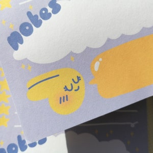 May include: A light purple notepad sheet featuring the word notes in blue, a large white cloud, yellow stars, and a yellow speech bubble next to a small yellow character with a smiling face and closed eyes, all printed on a matte paper surface.