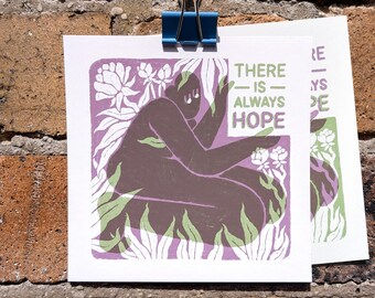 May include: A square art print featuring a dark brown figure kneeling amidst light green and white stylized flowers and foliage. The text "THERE IS ALWAYS HOPE" is printed in green on the right side. The print is held by a blue metal binder clip.